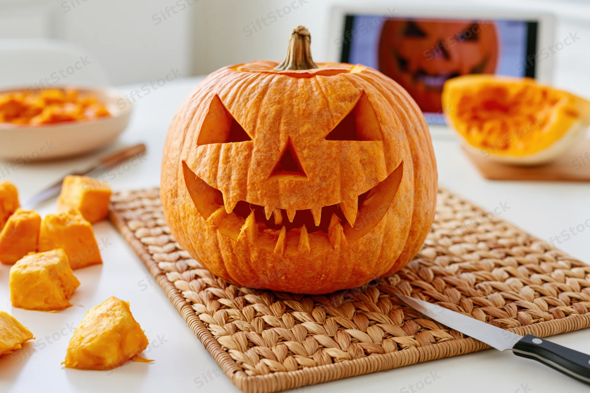 Carved pumpkin with triangular eyes and jagged teeth on woven mat in kitchen with knife and pumpkin pieces nearby