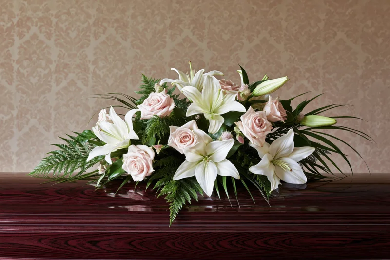 White lilies and pink roses arranged on a dark wooden coffin against patterned wallpaper