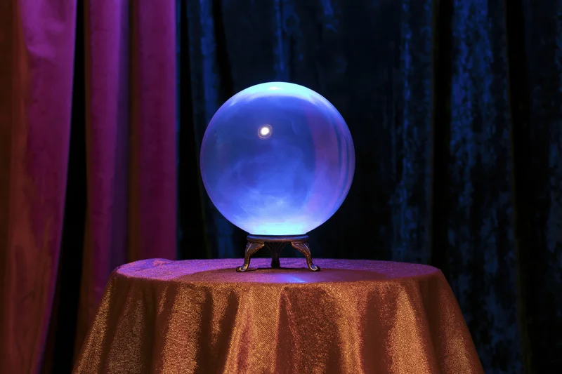 Glowing crystal ball on ornate metal stand atop table with draped fabric and curtains in background