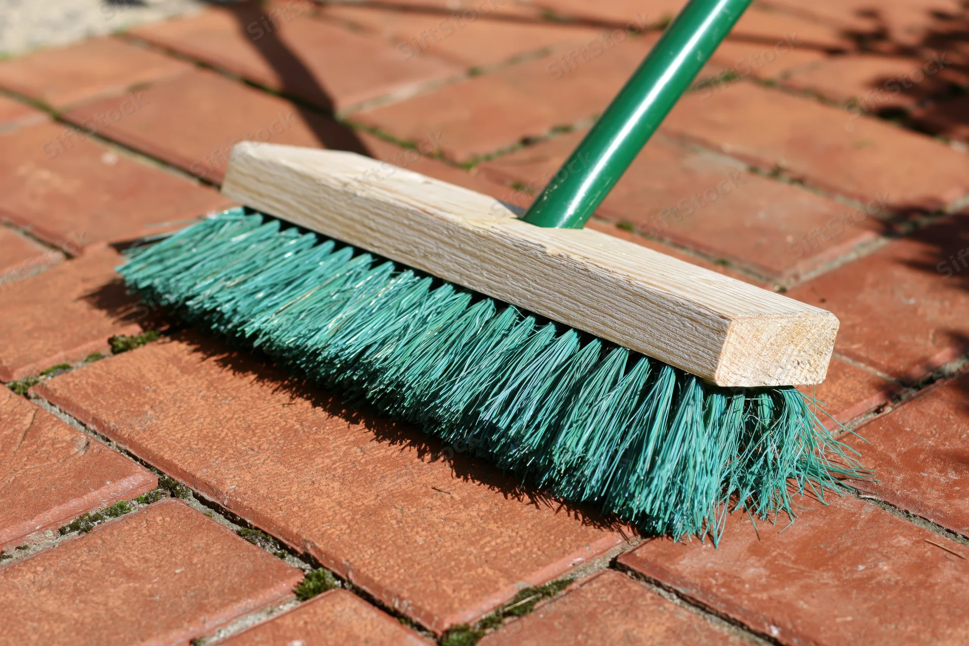 Green plastic-bristled push broom resting on red brick patio surface