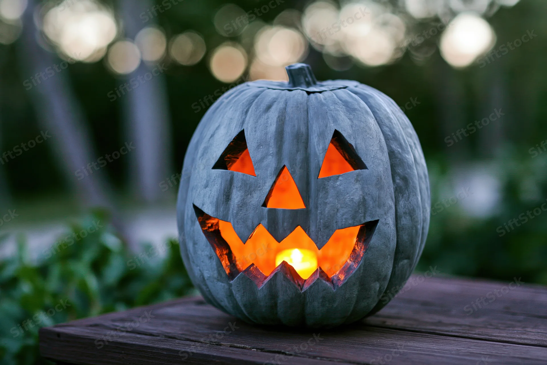 Grey painted carved pumpkin jack-o-lantern glowing with candlelight on wooden table outdoors at evening