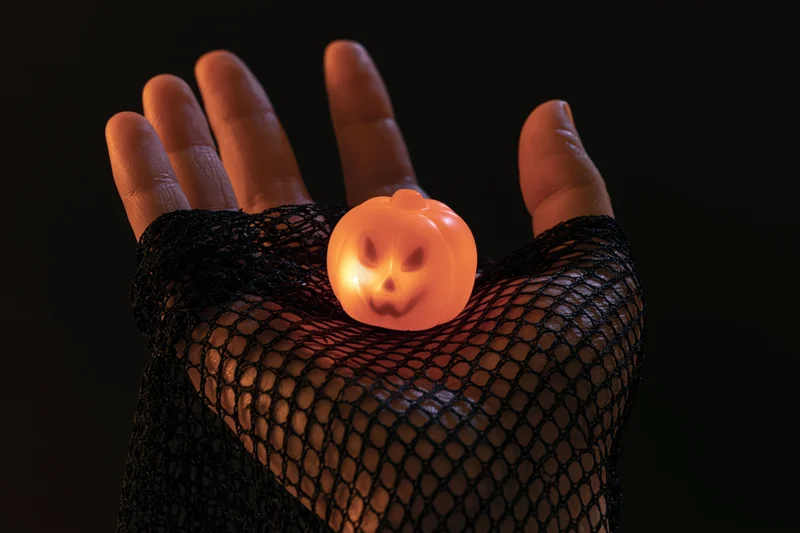 Hand in black fishnet glove cradling small glowing pumpkin against dark background