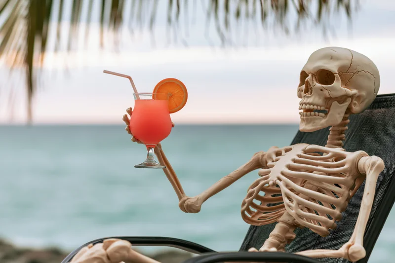 Skeleton reclining in beach chair holding cocktail by ocean at sunset
