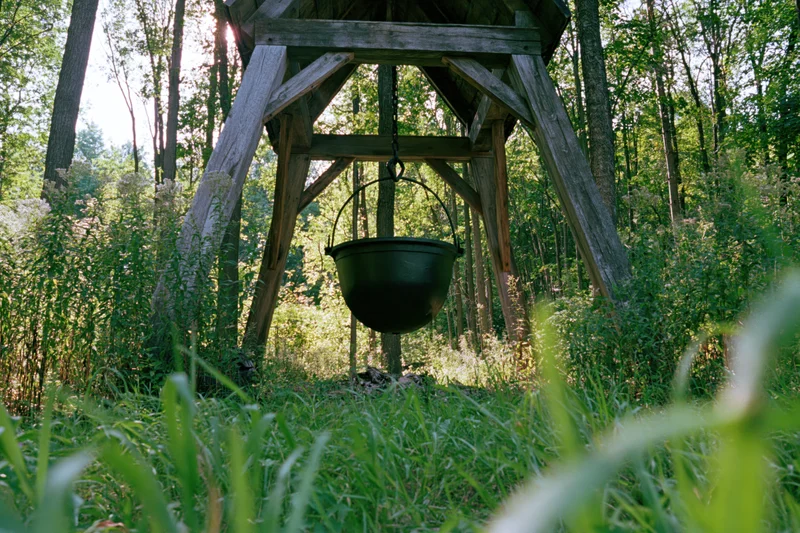 Black cast iron cauldron hanging from wooden frame in forested outdoor setting