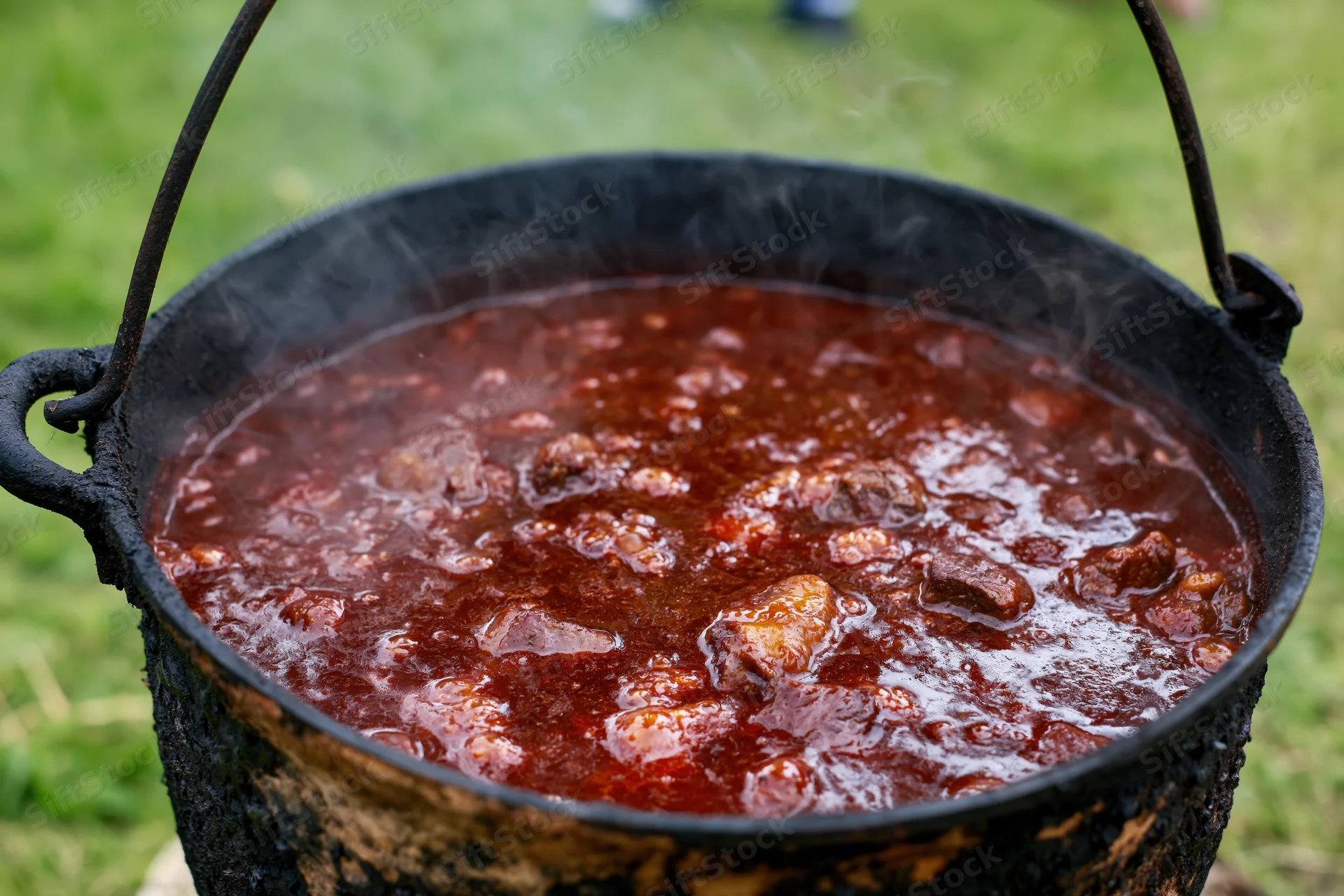 Large blackened cast iron cauldron simmering rich meat stew over an outdoor fire