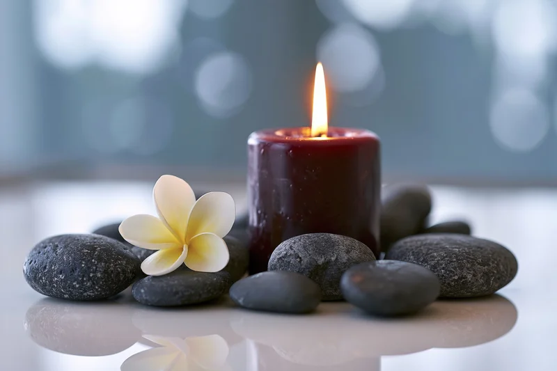 Lit burgundy pillar candle surrounded by smooth grey stones and a frangipani flower on a reflective surface