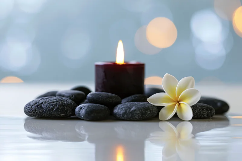 Lit burgundy candle surrounded by smooth black stones and a white frangipani flower on a reflective surface