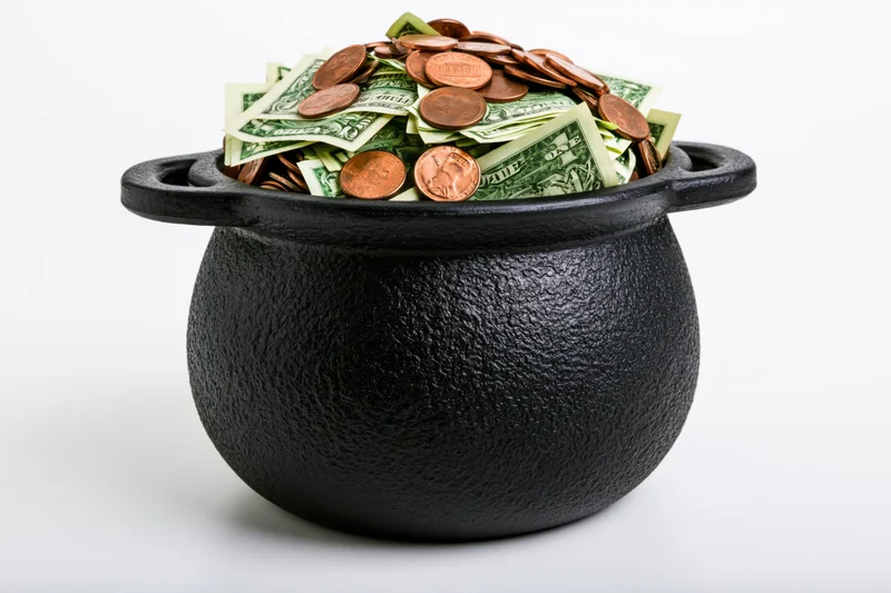 Black cast-iron cauldron overflowing with us dollar bills and coins on white background