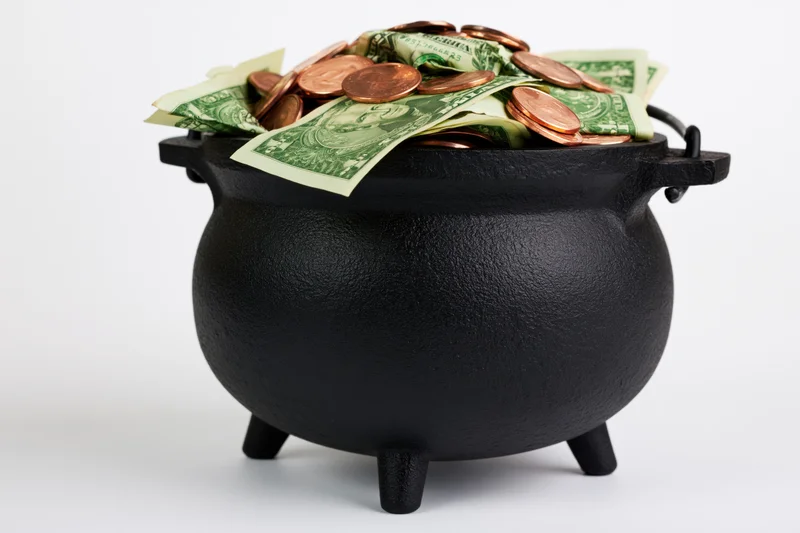 Black cast-iron cauldron overflowing with us dollar bills and coins on white background