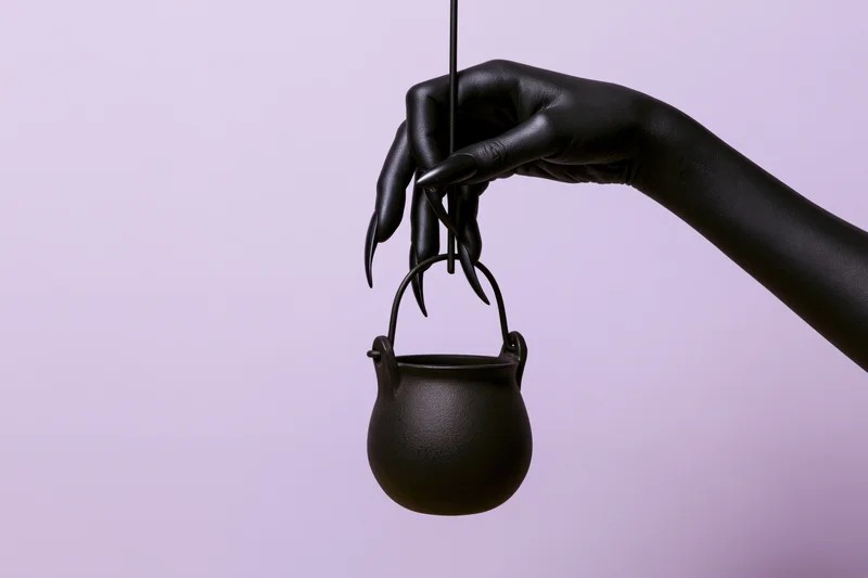 Black sculptural hand with clawed nails holding a small cauldron against a purple background