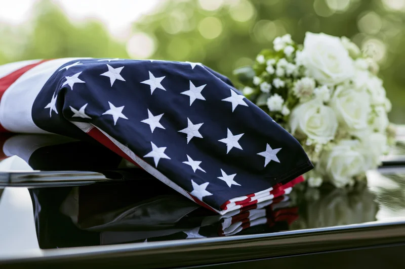 Folded american flag resting on a glossy black casket lid beside a bouquet of white roses