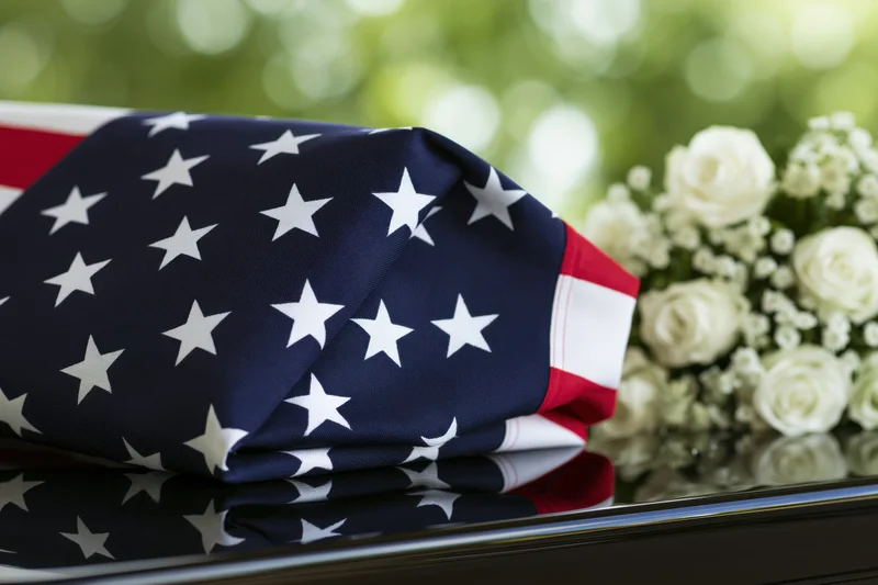Folded american flag resting on a glossy dark casket beside a bouquet of white roses
