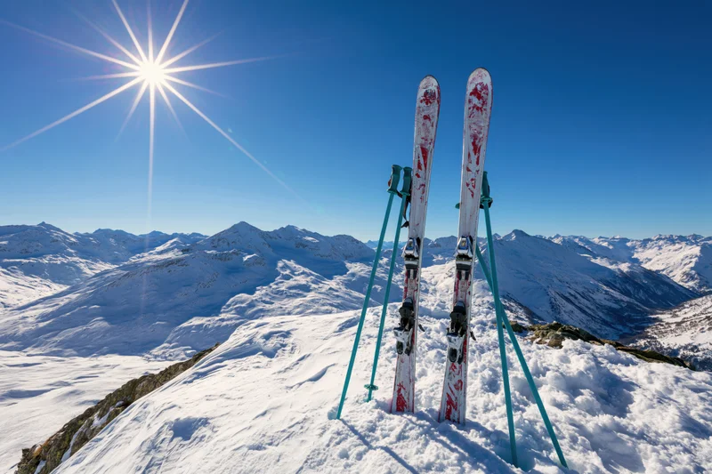 Pair of alpine skis and teal ski poles planted upright in snow on mountain summit under bright sun