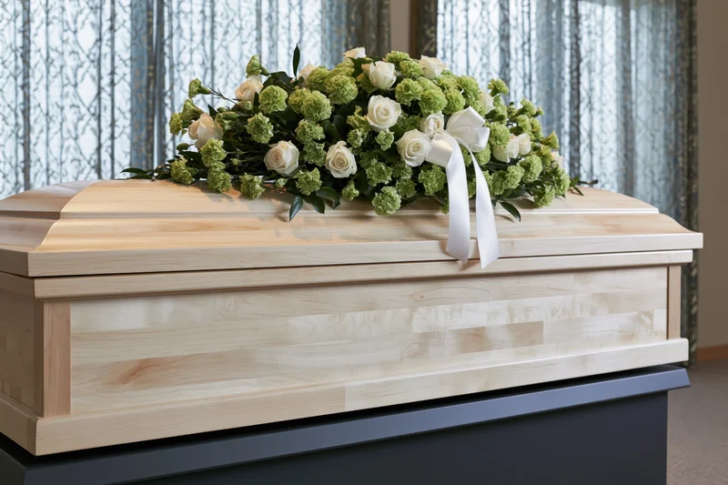 Pale birch wood coffin with white and green floral arrangement on dark bier indoors