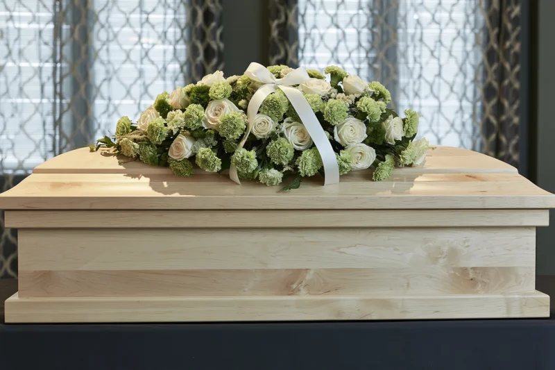 Pale birch wood coffin with white and green floral arrangement on dark bier near window with patterned curtains