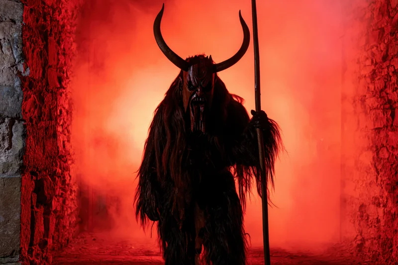 Krampus performer in horned mask and fur costume holding staff in stone corridor with red smoke