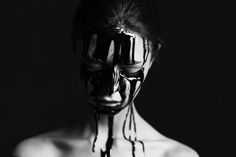 Woman with closed eyes and black paint dripping from her face and neck, bare shoulders, studio portrait