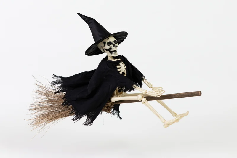 Skeleton wearing witch hat and black cape flying on broom against white background