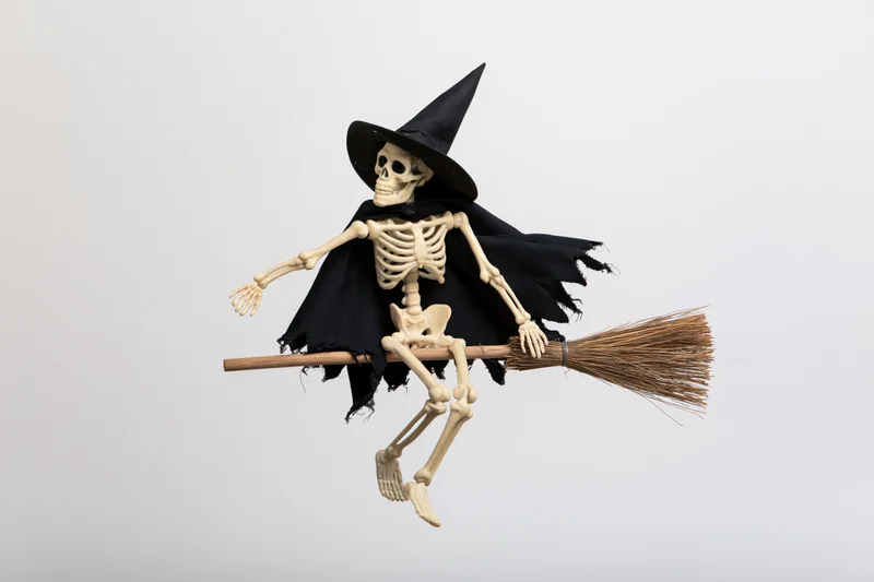 Skeleton wearing witch hat and cape flying on broom against white background
