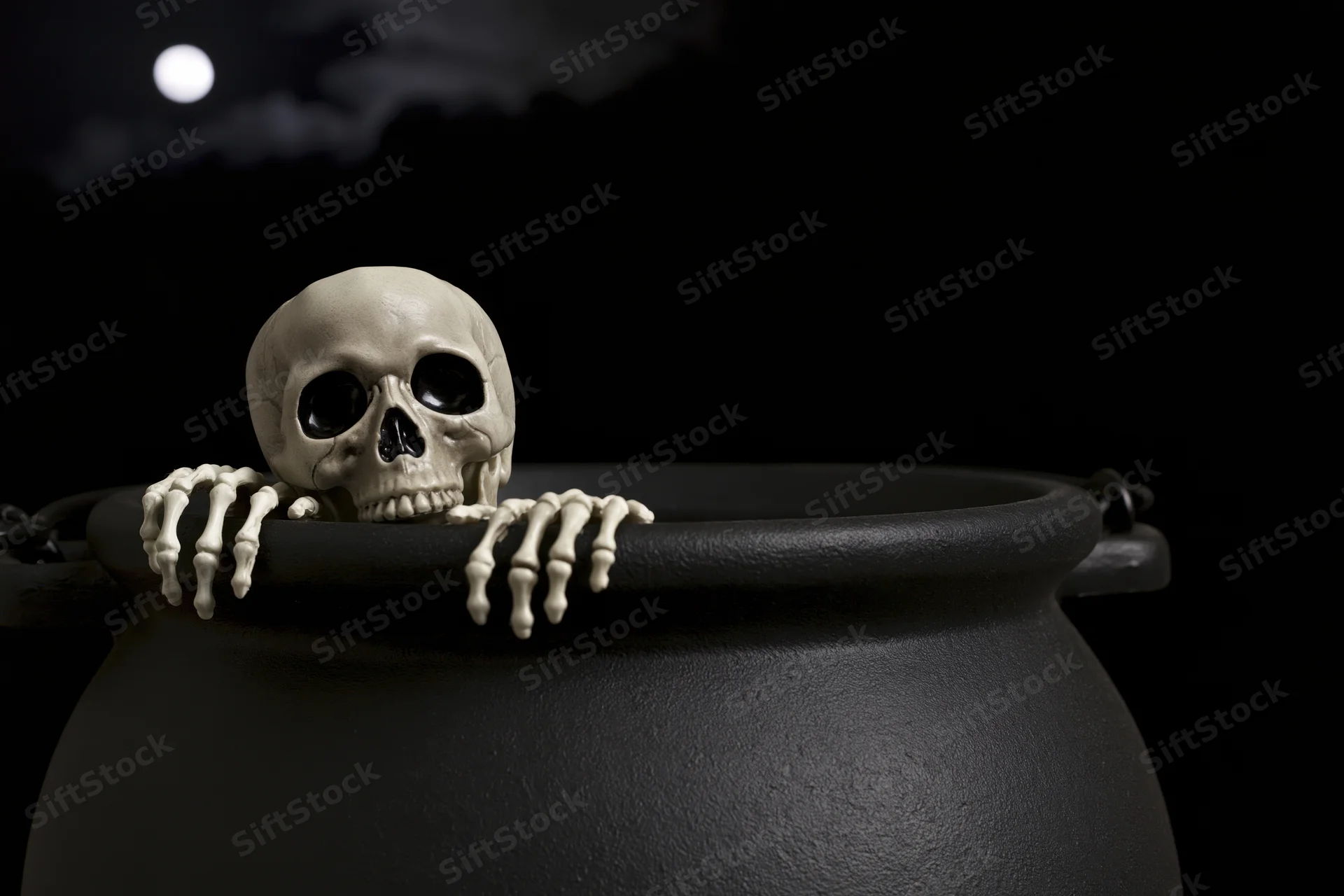 Plastic skeleton prop peering over black cauldron rim under moonlit night sky
