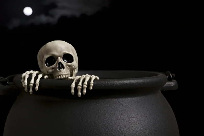 Plastic skeleton prop peering over black cauldron rim under moonlit night sky