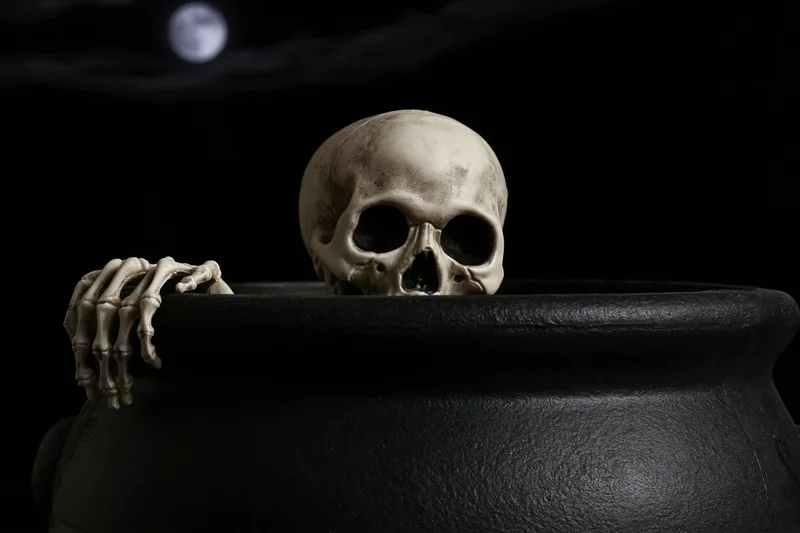 Plastic skeleton peering over black cauldron rim at night with full moon in dark sky