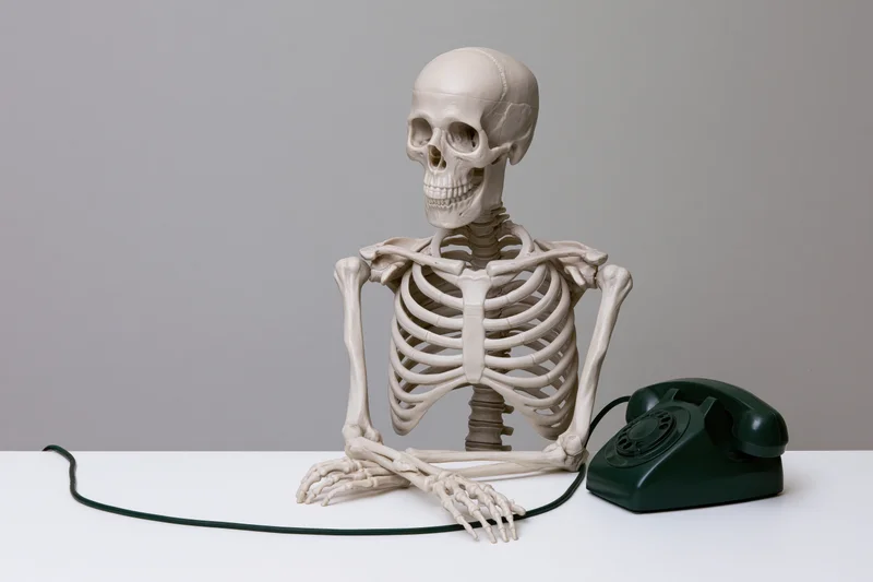 Plastic human skeleton model sitting upright at a white table beside a green rotary telephone