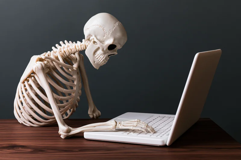 Plastic human skeleton model typing on laptop at wooden desk