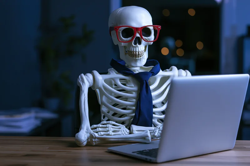 Skeleton wearing red glasses and blue tie working on laptop at desk