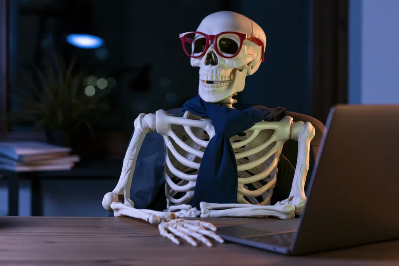 Plastic skeleton wearing red glasses and blue tie working on laptop at desk in dim office at night