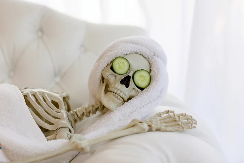 Plastic skeleton reclining on white tufted leather sofa with cucumber slices over eyes and towel wrapped around head