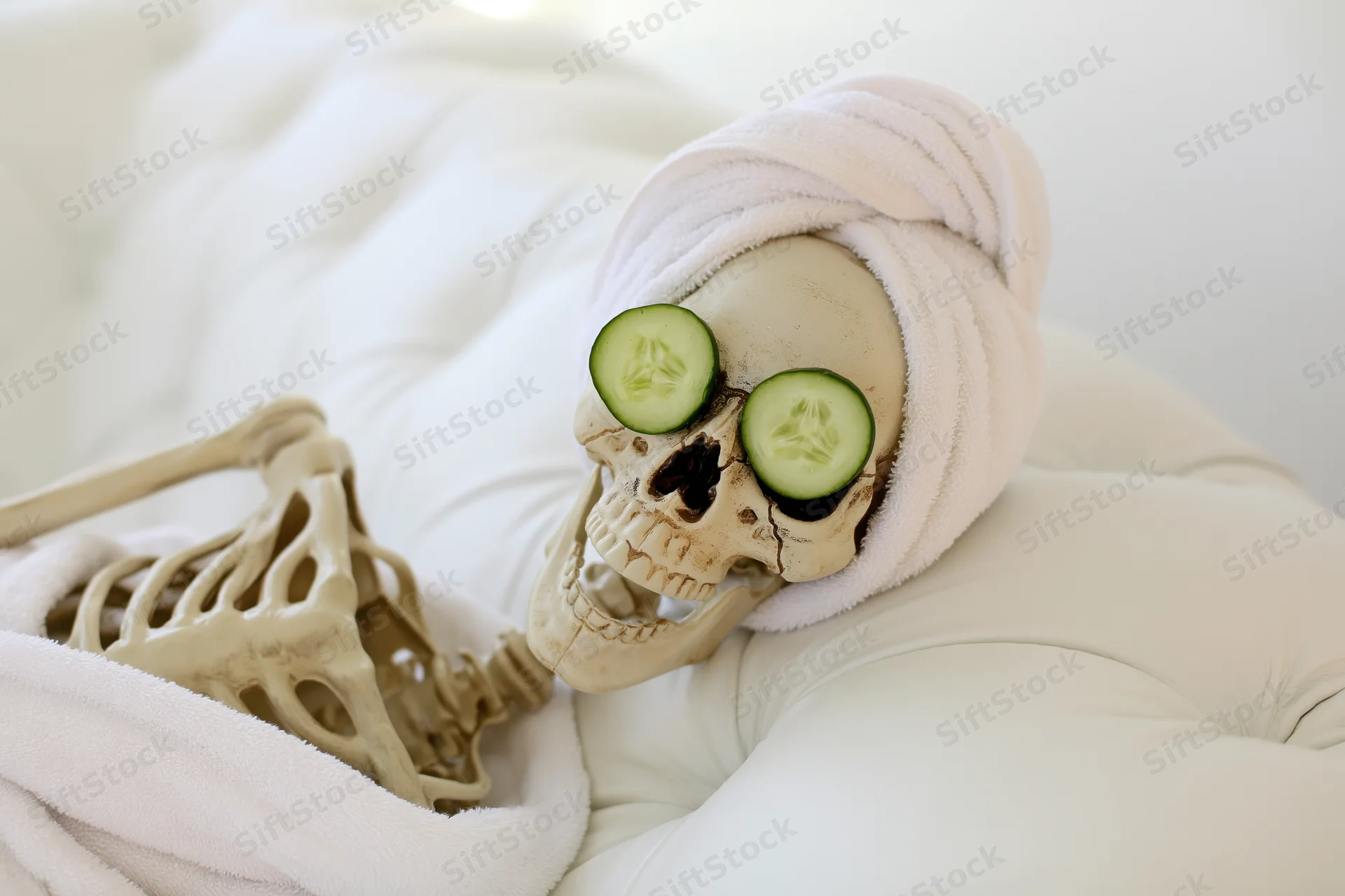 Plastic skeleton reclining on white tufted leather sofa with cucumber slices over eyes and towel wrapped around head
