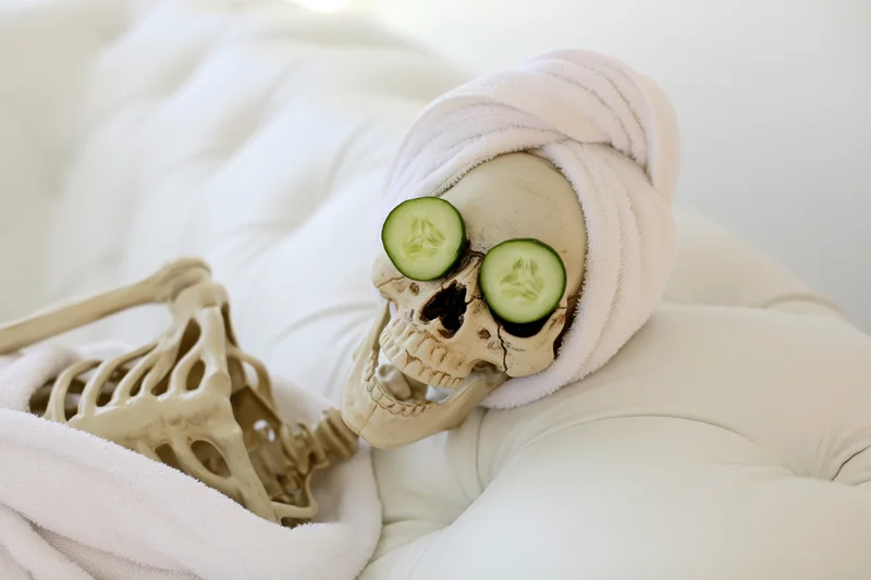 Plastic skeleton reclining on white tufted leather sofa with cucumber slices over eyes and towel wrapped around head