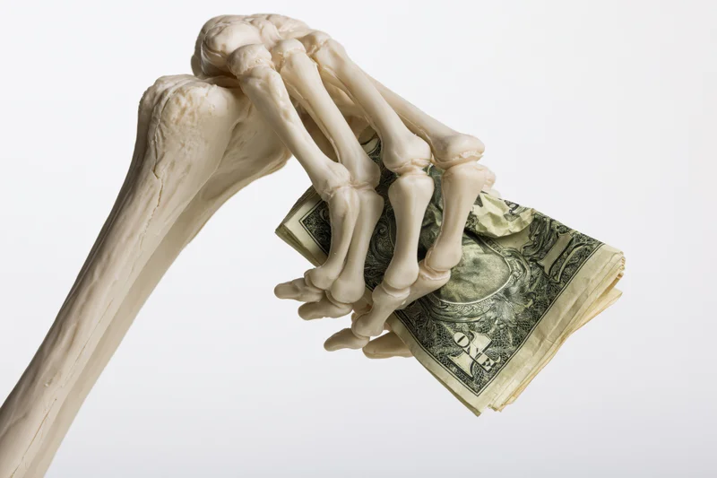 Skeleton hand gripping crumpled us dollar bills against white background