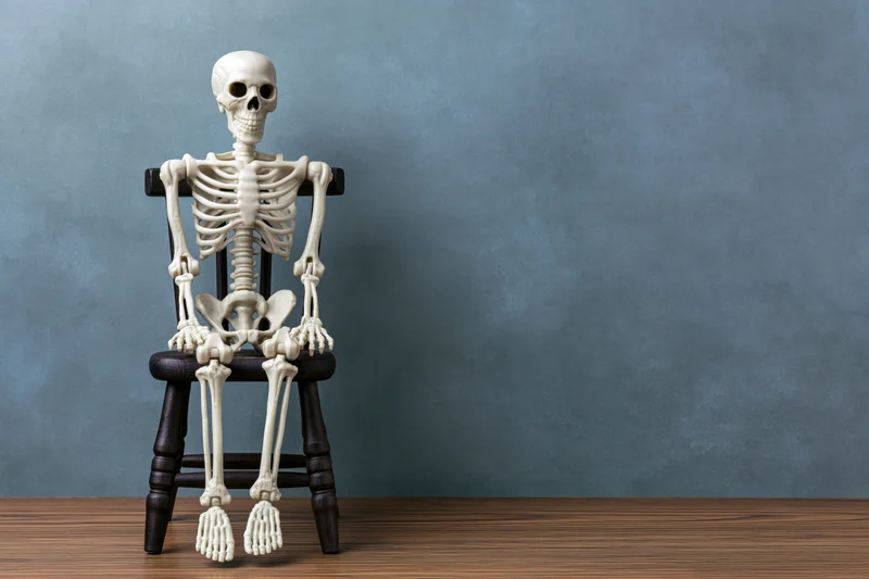Plastic toy skeleton sitting upright on a small wooden chair against a blue wall