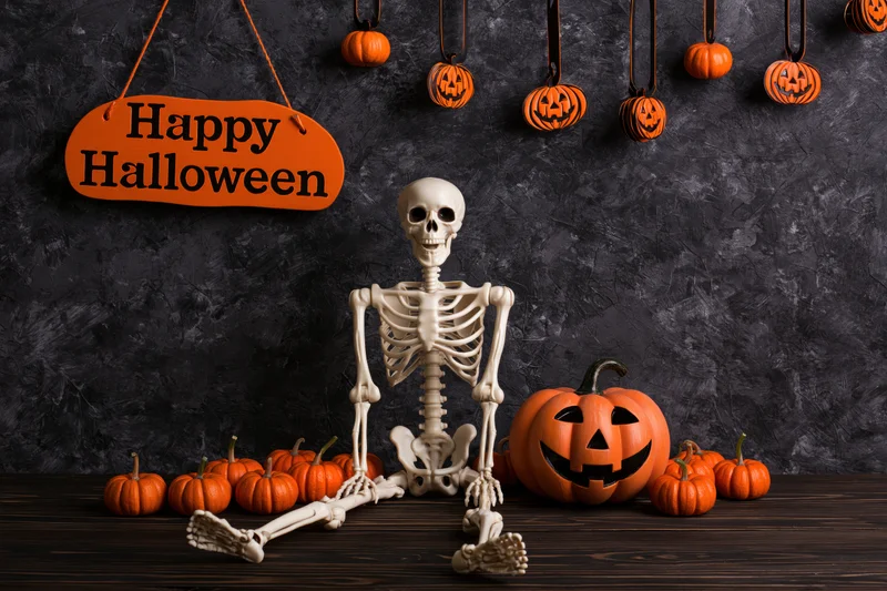 Plastic skeleton sitting upright on dark wooden table surrounded by carved pumpkins and hanging decorations for halloween