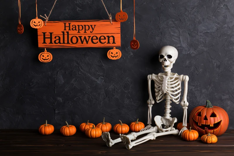 Plastic skeleton sitting upright on dark wooden surface surrounded by pumpkins and a hanging happy halloween sign