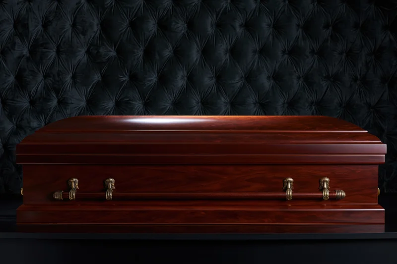 Polished mahogany casket with brass handles resting on a black tufted leather surface in a funeral home setting