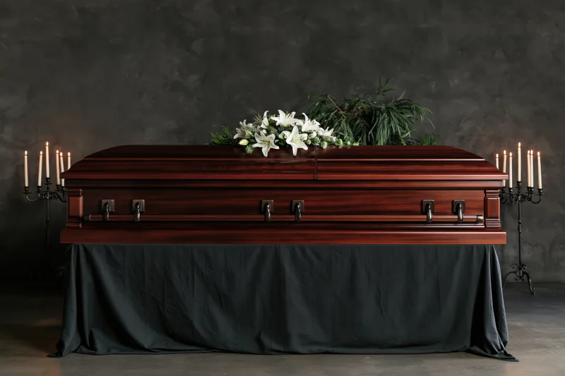 Polished mahogany casket adorned with white lilies and greenery, flanked by lit candles on candelabras, resting on a black-draped stand against a dark textured wall
