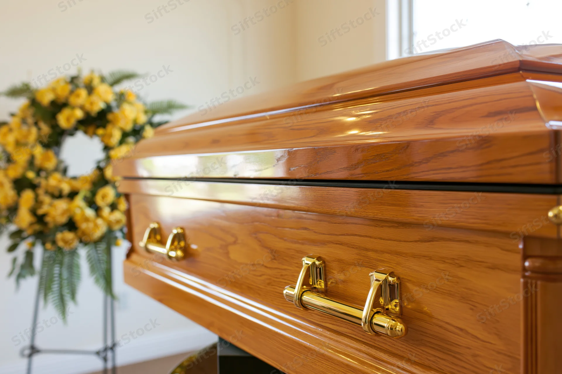 Polished wooden coffin with gold handles in a funeral home interior, floral wreath in background