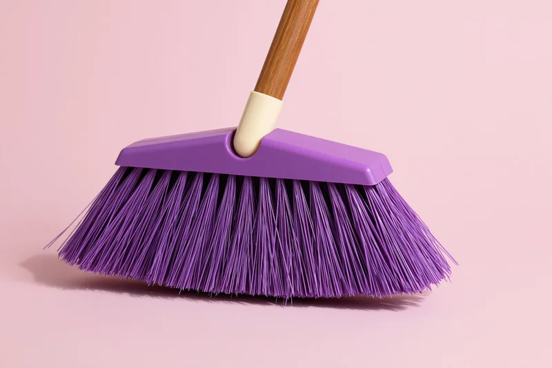 Purple plastic broom with dense synthetic bristles and wooden handle on pink background