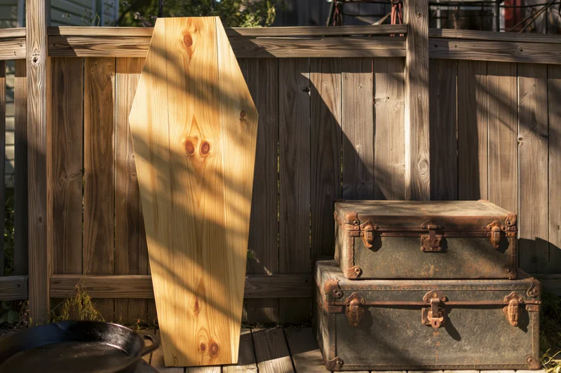 Raw pine coffin leaning upright against weathered wooden fence beside stacked vintage trunks in backyard