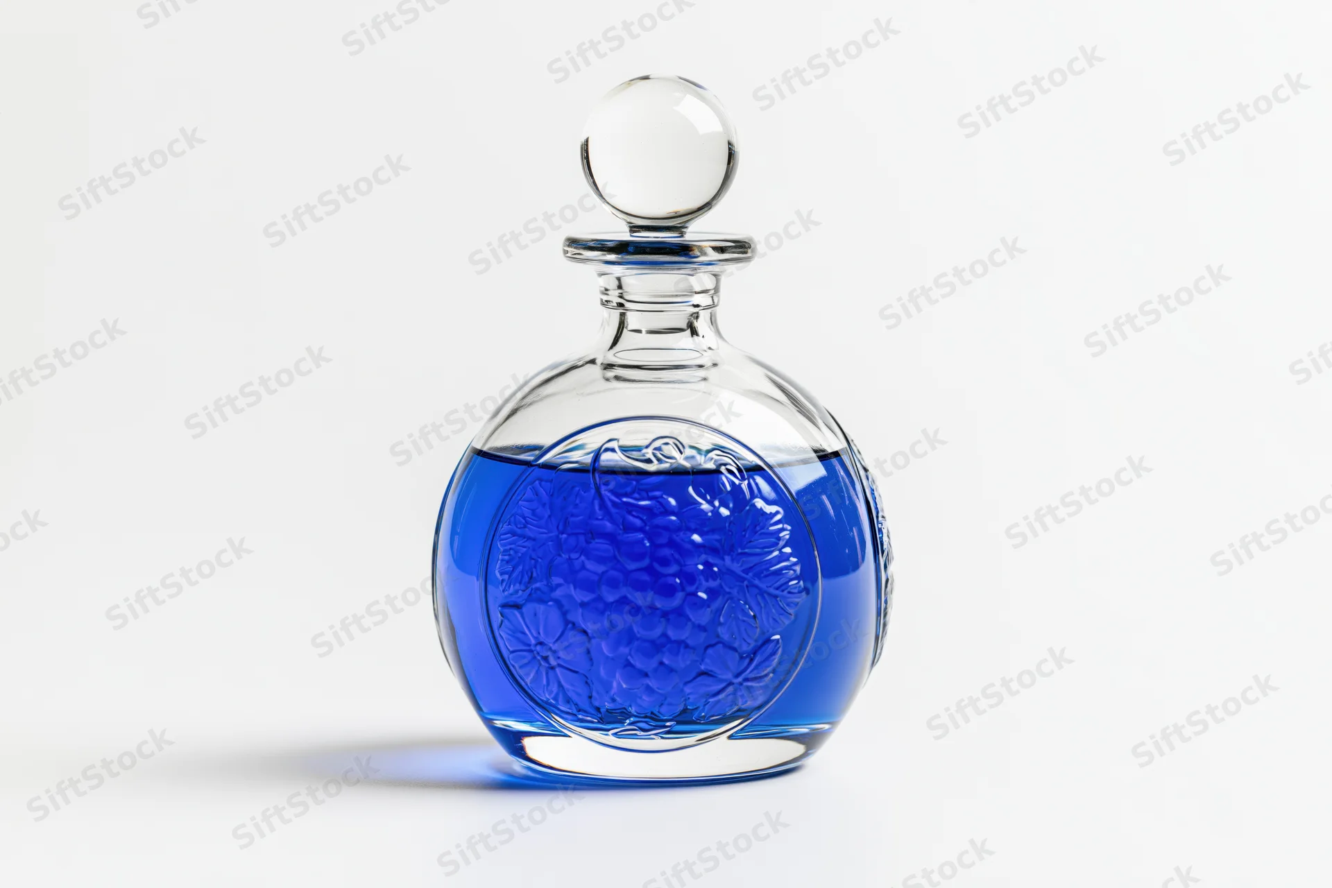 Round glass decanter with embossed floral pattern filled with blue liquid on white background