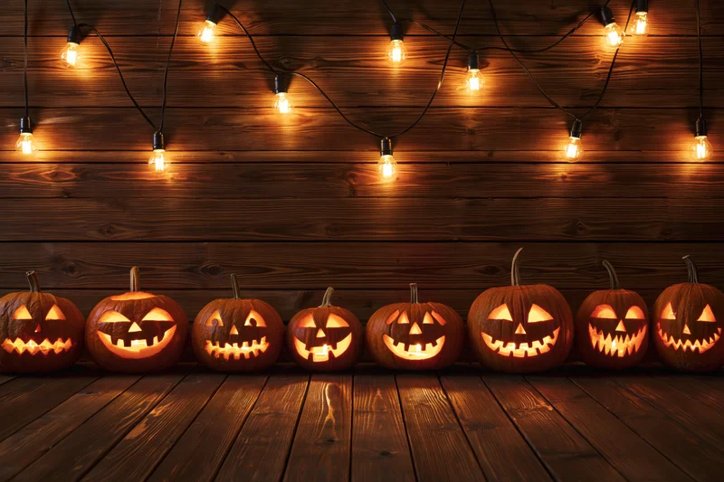 Row of carved jack-o-lanterns glowing on wooden floor beneath string lights