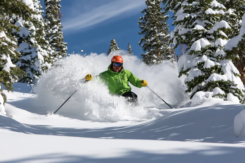 Skier in bright green jacket and orange helmet carving through deep powder snow on a mountain slope surrounded by snow-covered pine trees