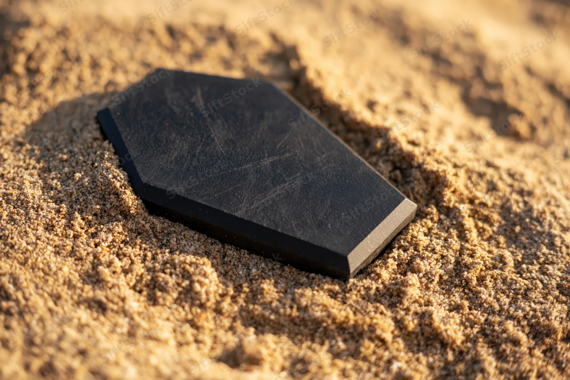 Black coffin-shaped object resting flat in coarse sand under warm sunlight