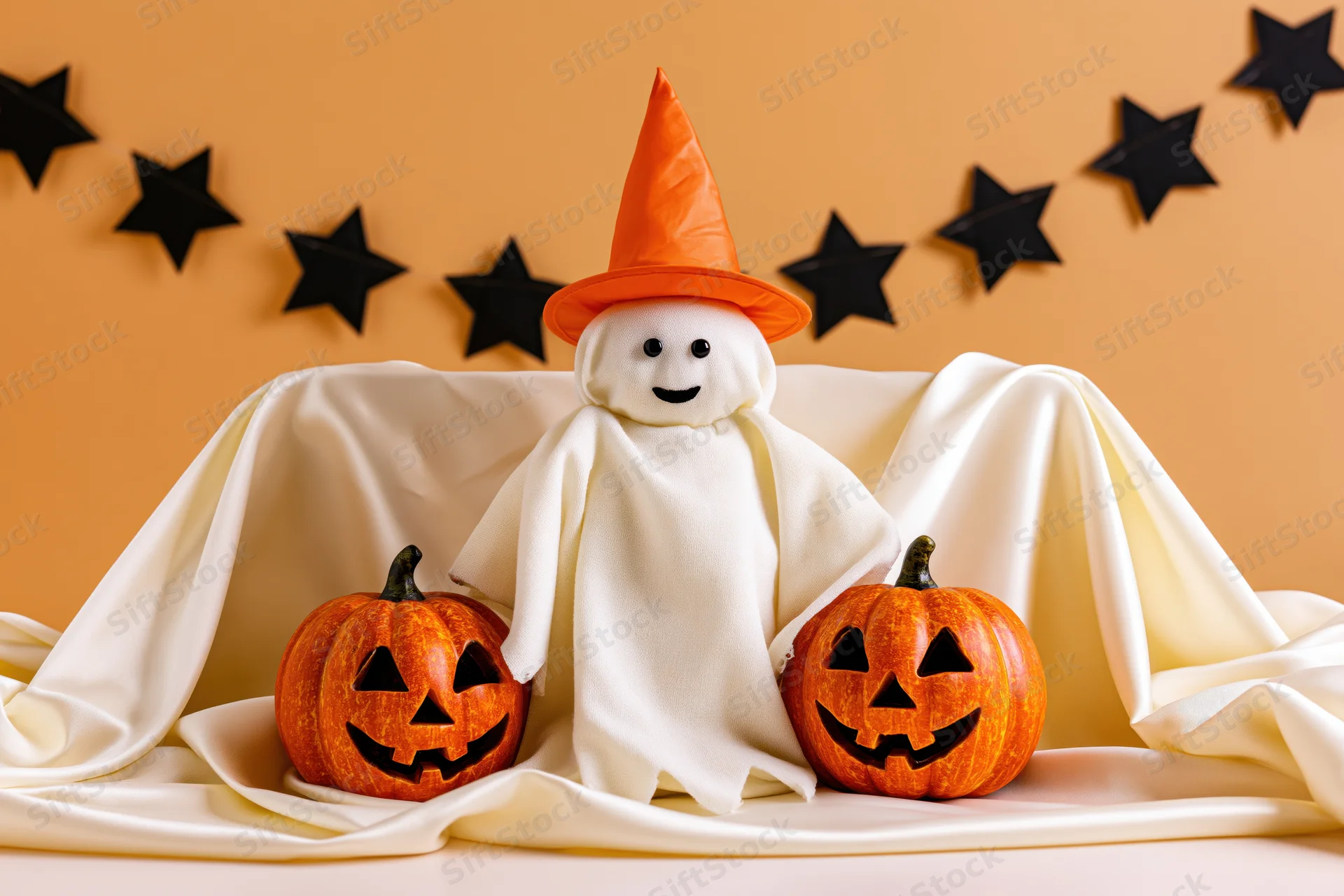 Small ghost costume figure with orange witch hat and jack-o-lanterns on draped fabric against orange wall with black star garland