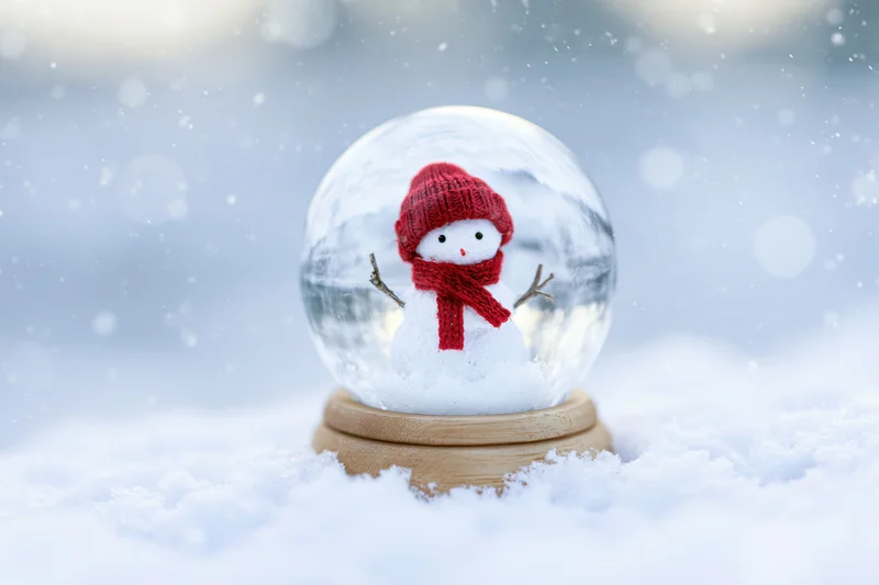 Snowman figure in red hat and scarf inside glass snowglobe resting on snow-covered ground outdoors