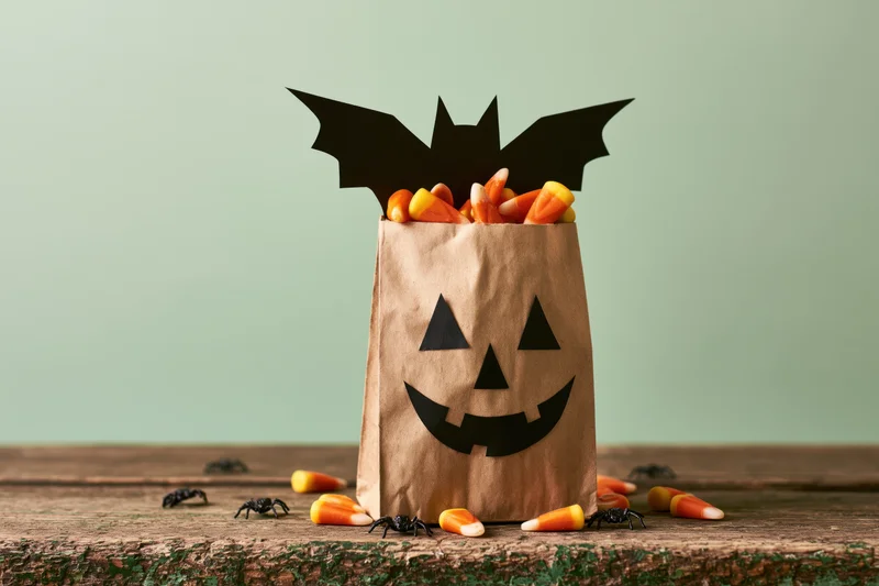 Decorated paper bag with jack-o-lantern face and bat wings filled with candy corn on wooden surface