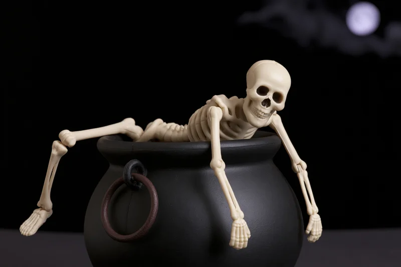 Plastic skeleton figurine draped over black cauldron against dark night sky with moon